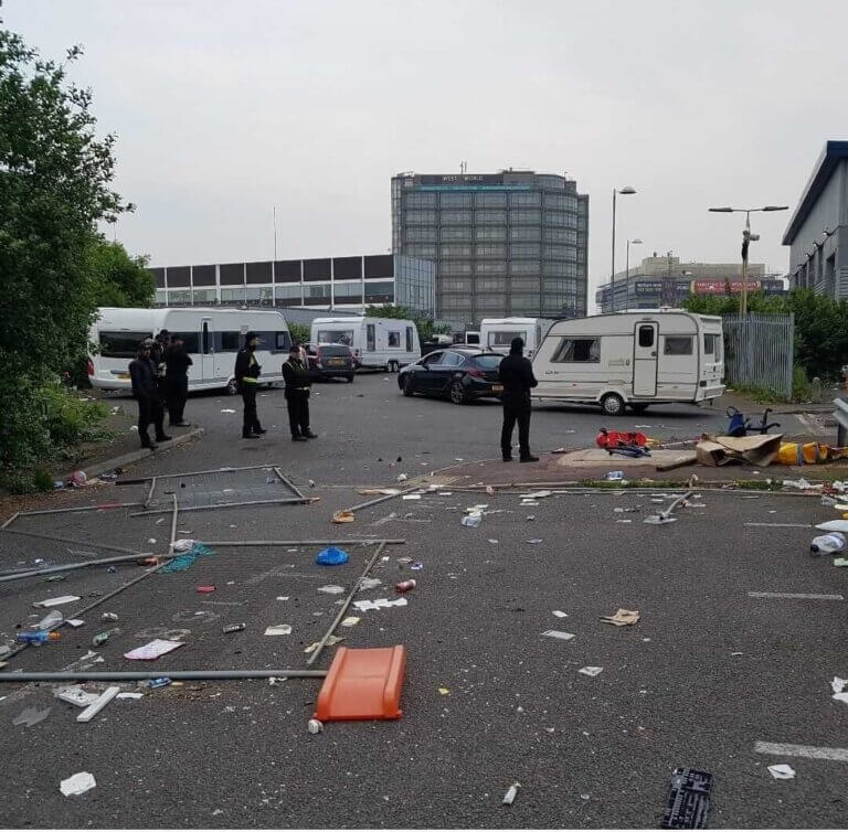 Enforcement officers managing an illegal traveller encampment in a littered commercial car park with caravans and debris.