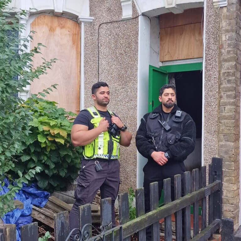 Two professional security enforcement officers standing guard in front of a boarded-up residential property.